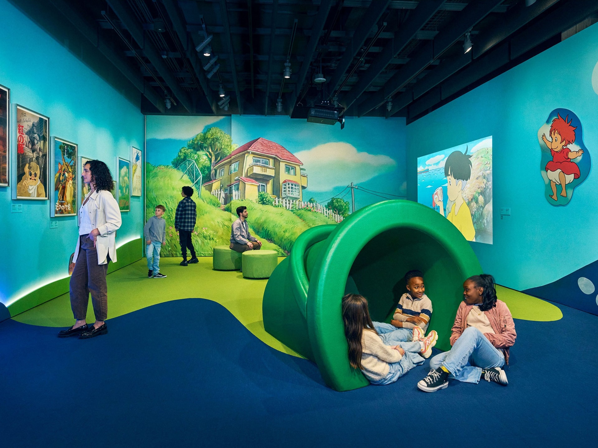 Visitors explore Ponyo (2008) artwork and immersive displays in the Ponyo exhibition at the Academy Museum of Motion Pictures, including a colorful seaside mural backdrop and a large green play structure where children sit and interact.